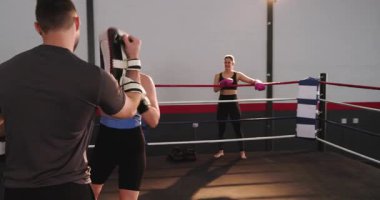 Koç, boks ringinde ulaşmak için odak yastığı tutuyor. Beyaz kadın yumruk-kroşe kombinasyonu yapıyor. Eğitim, fitness, atletizm, idman, güç, disiplin, hareket