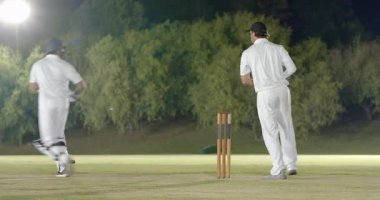 Bowling topu sahada, Hintli vurucu sopa sallıyor, beyaz kaleci antrenman için değiş tokuş yapıyor. Kriket, atletler, yarışma, takım çalışması, spor, açık hava, aksiyon