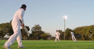 Kriket sopasıyla topa vurmak, farklı takım arkadaşları projektör kulesinin yakınında koşmak, kopyalama alanı. Atletik, takım çalışması, açık hava, sportmenlik, dinamik, aksiyon, atletizm