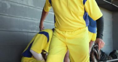 Yedek kulübesine giren, sarı formalı erkek beyzbol takım arkadaşları bankta selamlaşıp eldiven takıyorlar. Sporcular, yoldaşlık, takım çalışması, sportmenlik, günlük, açık hava, canlı