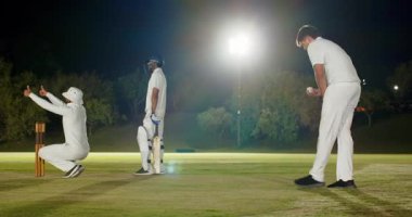 Kriket bovlingçisi, bekçinin topu durdurmak için baş parmağını kaldırmasından sonra projektörlerin altında bovling oynuyor. Spor, rekabet, atletizm, takım çalışması, açık hava, gece, aksiyon