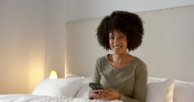 Afro-Amerikalı bir kadının akıllı telefonu dinleyip yatakta kitap okumasından sonra. Teknoloji, yaşam tarzı, modern, iletişim, rahatlama, konforlu, yerli