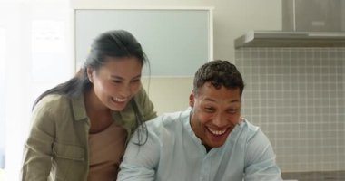 Asyalı kadın elini erkeğin omzuna koyuyor ve onu parlak mutfakta dizüstü bilgisayara yazmaya zorluyor. İşbirliği, samimiyet, modern, sıcak, takım çalışması, iletişim, yerli