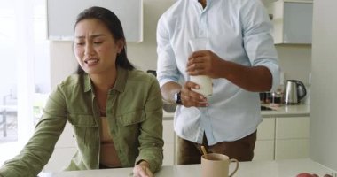 Asyalı çift sabah rutinini mutfak adasında paylaşıyor, adam tablet ekranı işaret ediyor ve süt döküyor. Samimiyet, bağlantı, teknoloji, yaşam tarzı, modern, sıcaklık, şefkat