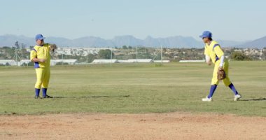Göğüs kafesinde eldiven tutuyor, erkek takım arkadaşları dış sahada topu atıyor ve yakalıyor, ayak çalışması yapıyor. Atletizm, antrenman, takım çalışması, hareket, sportmenlik, açık hava, rekabetçi