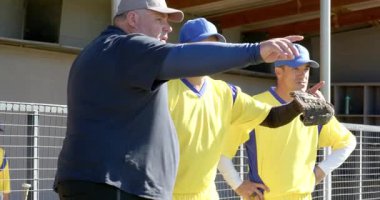 Sağa dönen erkek antrenör ve oyuncular yedek kulübesinde beyzbol eldivenleriyle saha pozisyonlarını özetliyorlar. Atletik, takım çalışması, akıl hocalığı, strateji, açık hava, canlı, aktif