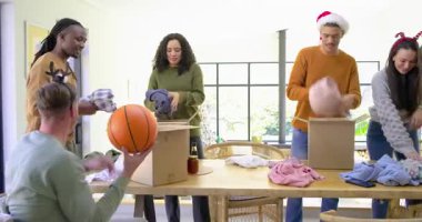 Çeşitli arkadaşlar masadaki kutulara tatil kıyafetleri ve kavanozlar dolduruyor, oturan adam basketbol topluyor. İşbirliği, kutlama, şenlik, sosyal dinamik, neşeli, toplum, kırsal