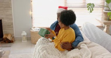 Afro-Amerikalı çift yeşil hediye kutusu tutuyorlar açılış hediyesi olarak kurdeleyi açıyorlar ve kanepede sarılıyorlar. Romantizm, samimiyet, samimiyet, kutlama, sıcaklık, sevgi, evcil