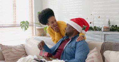 Noel Baba şapkalı ve sarı kazaklı farklı bir çift kanepenin üzerinde çözülen kurdeleyi açıyor. Romantik, neşeli, rahat, kutlama, samimiyet, neşe, tatil