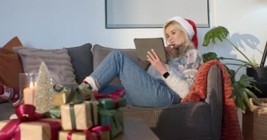 Oturma odasındaki kanepede tableti kaldıran kadın hediye ve mumların arasında kayıyor. Kopya alanı. Şen, rahat, kutlama, tatil, boş zaman, iç mekan, konfor