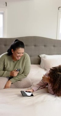 Dikey video: Anne ve kızı yatak başlığında telefon ve tablet değiş tokuşu yaparken görülüyor. Aile, bağ, teknoloji, rahatlık, konfor, samimiyet, dikey video