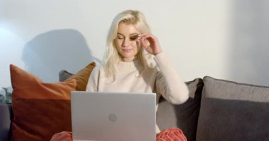 Kanepeye kurulmuş gümüş dizüstü bilgisayarı açan, kaşlarını okşayan ve çalışırken gülümseyen bir kadın. Konsantrasyon, rahatlama, rahat, konforlu, teknoloji, üretkenlik, konfor