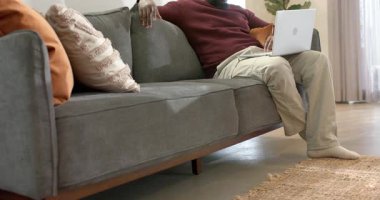 Kanepede oturuyor, Afro-Amerikalı bir adam dizüstü bilgisayar yazıyor, ekranda kayıyor ve gülüyor, boşluğu kopyalıyor. Günlük, ev ofisi, rahat, rahatlama, teknoloji, yaşam tarzı, modern