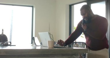 Mutfakta Afro-Amerikalı bir adam akıllı telefon alıyor. Laptop ve kupa kullanarak, kopyalama alanı kullanarak. Üretkenlik, iletişim, modern, iç mekan, yaşam tarzı, teknoloji, çalışma