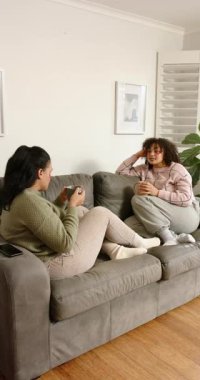 Dikey video: Göz kremi süren, fincanları yudumlayan, gri koltukta sohbet eden çeşitli bayan arkadaşlar. Rahat, dostluk, rahatlama, cilt bakımı, yaşam tarzı, ev içi, dikey video