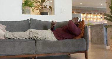 Kanepeye uzanmış, portakal rengi yastıklı bir adam içeri giriyor. Telefonları dinliyor ve kulaklıkları ayarlıyor. Rahatlama, rahatlama, teknoloji, yaşam tarzı, modern, konfor, huzur