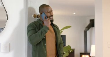 Afro-Amerikalı adam akıllı telefonlar alıyor, başını sallıyor, sırıtıyor ve gülüyor oturma odasında. Yaşam tarzı, rahatlama, modern, rahat, iletişim, sükunet, iç mekan