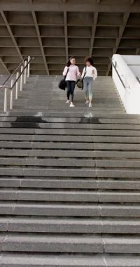 Dikey video: Karşılıklı bakış, Asyalı ve Afrikalı Amerikalı arkadaşlar çantalarla merdivenlerden iniyorlar. Kentsel, dostluk, yoldaşlık, aktivite, gezinti, modern, dikey video