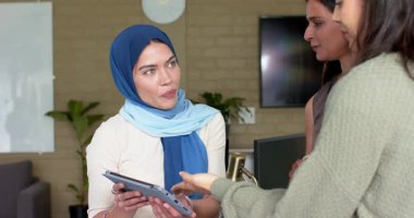 Tablet tesettürlü kadına doğru hareket eden yeşil kazaklı kadın demo çalıyor ve tezgahta takım izliyor. İşbirliği, takım çalışması, dijital, modern, profesyonel, çeşitlilik, yenilik