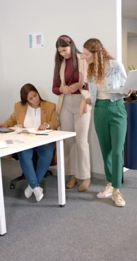 Dikey video: Çeşitli bayan takımları masa başında toplayıp notlar almak ve dizüstü bilgisayarı işaret etmek. İşbirliği, takım çalışması, profesyonellik, modernlik, iş kadınları, beyin fırtınası, dikey video