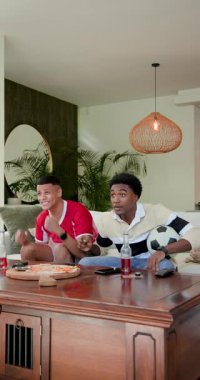 Dikey video: Kanepede top tutan çeşitli erkek arkadaşlar oturma odasındaki kaleye tepki veriyorlar. Kutlama, yoldaşlık, eğlence, modern, yaşam tarzı, neşe, dikey video