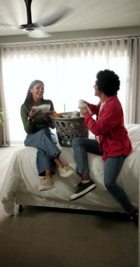 Dikey video: Odaya giren çeşitli arkadaşlar, temiz kıyafetler için çamaşır sepetinden çamaşır katlıyorlar. Günlük, arkadaşlık, yerli, yaşam tarzı, uyum, dinlenme, dikey video