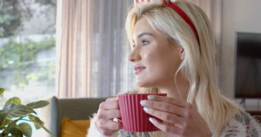 Pencereden süzülen güneş ışığı kırmızı kupayı tutan ve yavaşça kanepede yudumlayan kadına ilham veriyor. Rahat, rahat, sıcak, evcil, rahat, yaşam tarzı, minimalist