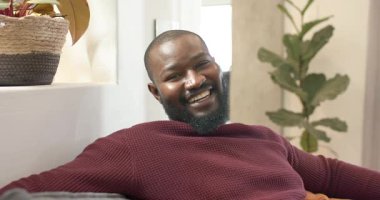 Afro-Amerikalı orta yaşlı bir adam kamera dışı sohbete tepki veriyor. Evdeki kanepede sırıtarak arkana yaslanıyor. Rahatlama, rahatlama, rahatlama, ev dekorasyonu, çağdaş, yaşam tarzı, gündelik