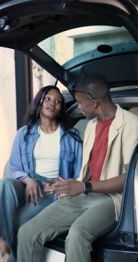 Dikey video: Afro-Amerikan çift akıllı telefon kullanarak araba yolculuğunun bagajında oturuyor. Macera, seyahat, boş zaman, açık hava, yaşam tarzı, modern, dikey video