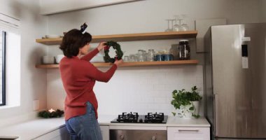 Mutfağa çelenk taşıyarak giren kadın, çelengi rafın altına koyuyor ve dekorasyon için ışıkları örtüyor. Şen, rahat, iç mekan, sade, sıcak, aydınlık, mevsimlik