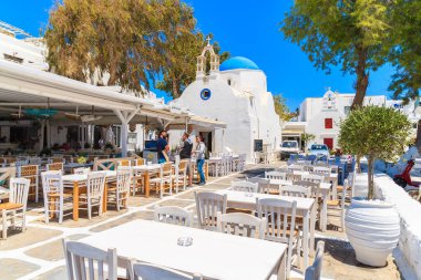 MYKONOS TOWN, GREECE - 16 Mayıs 2016: Yunanistan 'ın Mykonos kentinde beyaz bir kilise önünde tipik bir meyhanede sandalye ve masalarla konuşan Yunanlılar.