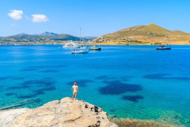 Genç kadın turist kayaların üzerinde duruyor ve Yunanistan 'ın Paros adasında yelkenli teknelerle mavi denize bakıyor.