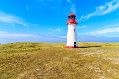 Kum tepesindeki deniz feneri mavi gökyüzüne karşı. Almanya 'nın List köyü yakınlarındaki Sylt Adası' nın kuzey sahilinde beyaz bulutlar.