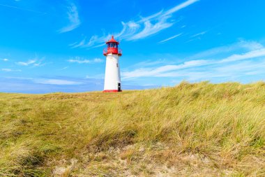 Kum tepesindeki deniz feneri mavi gökyüzüne karşı. Almanya 'nın List köyü yakınlarındaki Sylt Adası' nın kuzey sahilinde beyaz bulutlar.