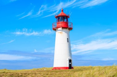 Kum tepesindeki deniz feneri mavi gökyüzüne karşı. Almanya 'nın List köyü yakınlarındaki Sylt Adası' nın kuzey sahilinde beyaz bulutlar.
