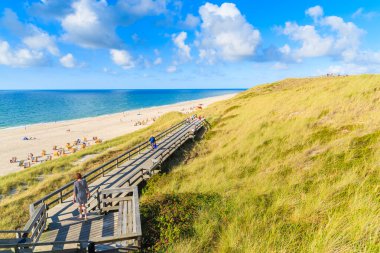Kuzey Denizi kıyısında ahşap bir yolda yürüyen kadın turist ve Almanya 'nın Sylt Adası' ndaki Wenningstedt köyü yakınlarındaki güzel plaj manzarası