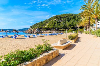 Güneşli yaz gününde İspanya 'nın İbiza adasında, Cala San Vicente körfezinde kumlu sahil boyunca şemsiyeli ve güneşli yataklı gezinti.