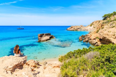 Kimliği belirsiz genç bir kadın bir kayanın üzerinde oturuyor ve İbiza, İspanya 'daki güzel Cala Tarida Körfezi' ne bakıyor.
