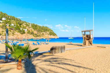 Güneşli yaz gününde İspanya 'nın İbiza adasında Cala San Vicente koyunda kumlu sahilde cankurtaran kulesi ve şemsiyeli güneşli yataklar.