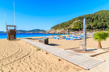 Güneşli yaz gününde Cala San Vicente koyunda şemsiyeli ve güneşli yataklı kumlu plaja giden yol, İbiza Adası, İspanya