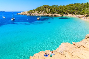 Tanımlanamayan iki genç kadın, İspanya 'nın İbiza adasında, Cala Salada koyunda kristal berraklığında turkuaz deniz suyuyla güneşleniyor.