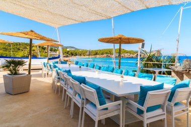 CALA NOVA BEACH, IBIZA ADASI - 20 Mayıs 2017: Cala Nova sahilindeki güzel deniz manzaralı restoran terasında beyaz masa ve mavi yastıklı sandalyeler, İbiza Adası, İspanya.