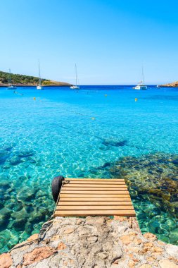 Ahşap iskele ve mavi deniz manzarası. Uzaklarda yelkenli tekneleri var. Cala Portinatx Körfezi, İbiza Adası, İspanya.