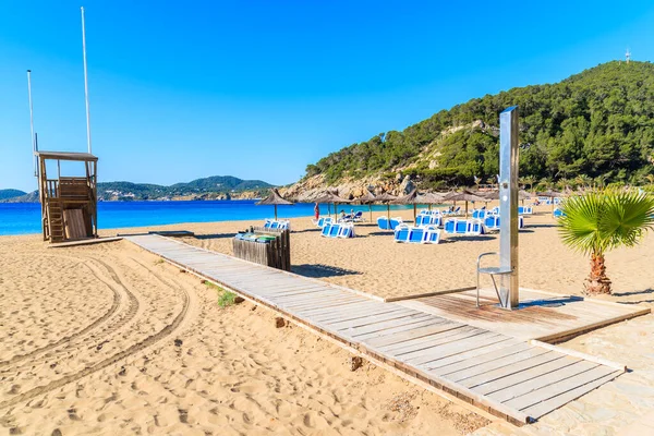 Güneşli yaz gününde Cala San Vicente koyunda şemsiyeli ve güneşli yataklı kumlu plaja giden yol, İbiza Adası, İspanya