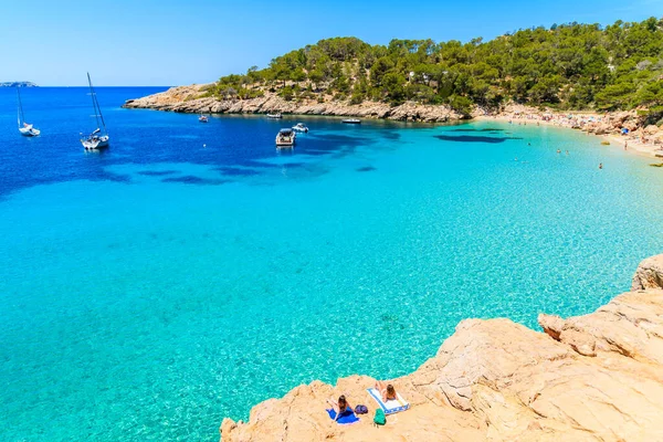 Tanımlanamayan iki genç kadın, İspanya 'nın İbiza adasında, Cala Salada koyunda kristal berraklığında turkuaz deniz suyuyla güneşleniyor.
