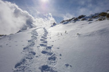 Bir dağ sırtındaki el değmemiş beyaz kar yığınına kazınmış derin ayak izlerinden oluşan çarpıcı bir perspektif.