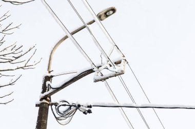 Bir sokak lambası ve arka planda karla kaplı bir sürü kablo. Gri gökyüzü ve donmuş ağaç.