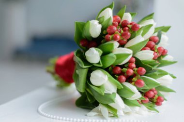 Düğün hazırlıkları: zarif bir buket beyaz çiçek, kırmızı böğürtlen ve yeşil yapraklar inci bir kolyenin yanındaki beyaz masada duruyor.