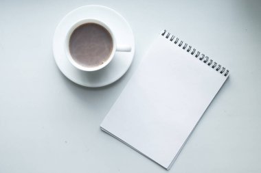 Bir fincan taze kahve ve kahverengi bir masanın üzerinde mavi ışıklı beyaz bir defter.