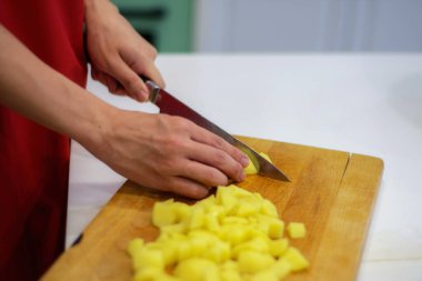 Kırmızı önlüklü bir adam büyük bir bıçakla tahta bir tahtanın üzerinde sarı patatesler keser.